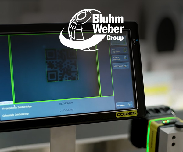 컨베이어 벨트 측면에 장착된 Cognex 머신 비전 시스템 옆, 화면 위 QR 코드 이미지 옆에 Bluhm Weber 로고가 보입니다.