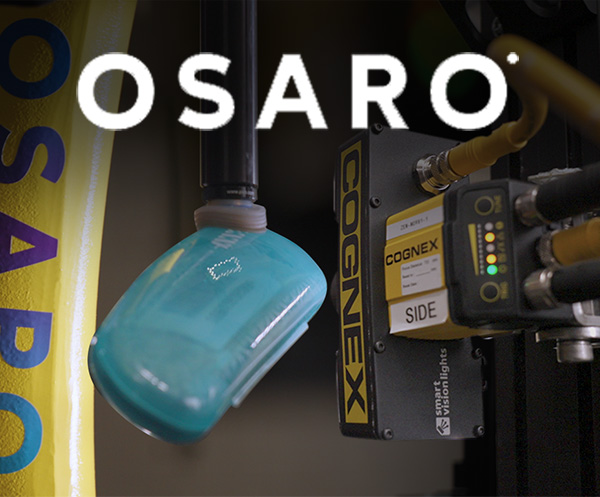 Das Osaro-Logo wird über einem Roboterarm gezeigt, der ein Brillenetui aufnimmt; ein Barcode-Lesegerät von Cognex ist an der Seite angebracht und erfasst den Barcode.